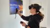 Eine Frau steht mit VR-Brille und 2 Controllern in den Händen vor einem Bildschirm an der Wand.